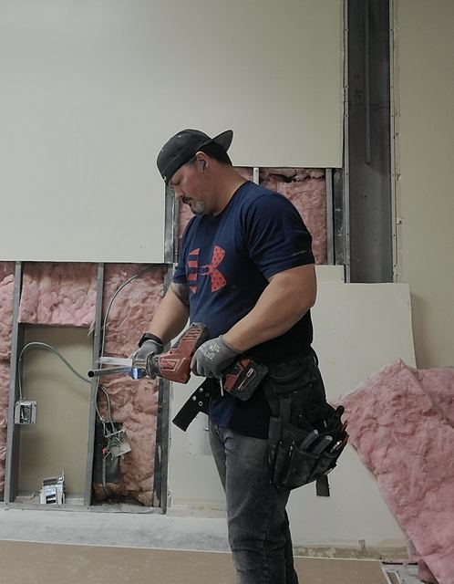 Parker Enterprises electrician cutting conduit with a reciprocating saw at an open wall with MC cable and electrical boxes